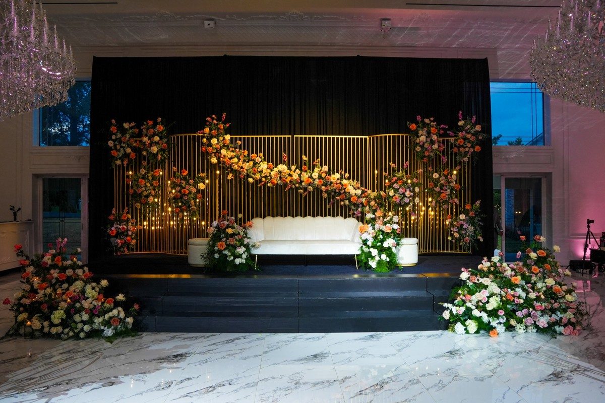 A stage with a gold slatted backdrop, coral and pink florals and an ivory sofa, designed by New Era Events