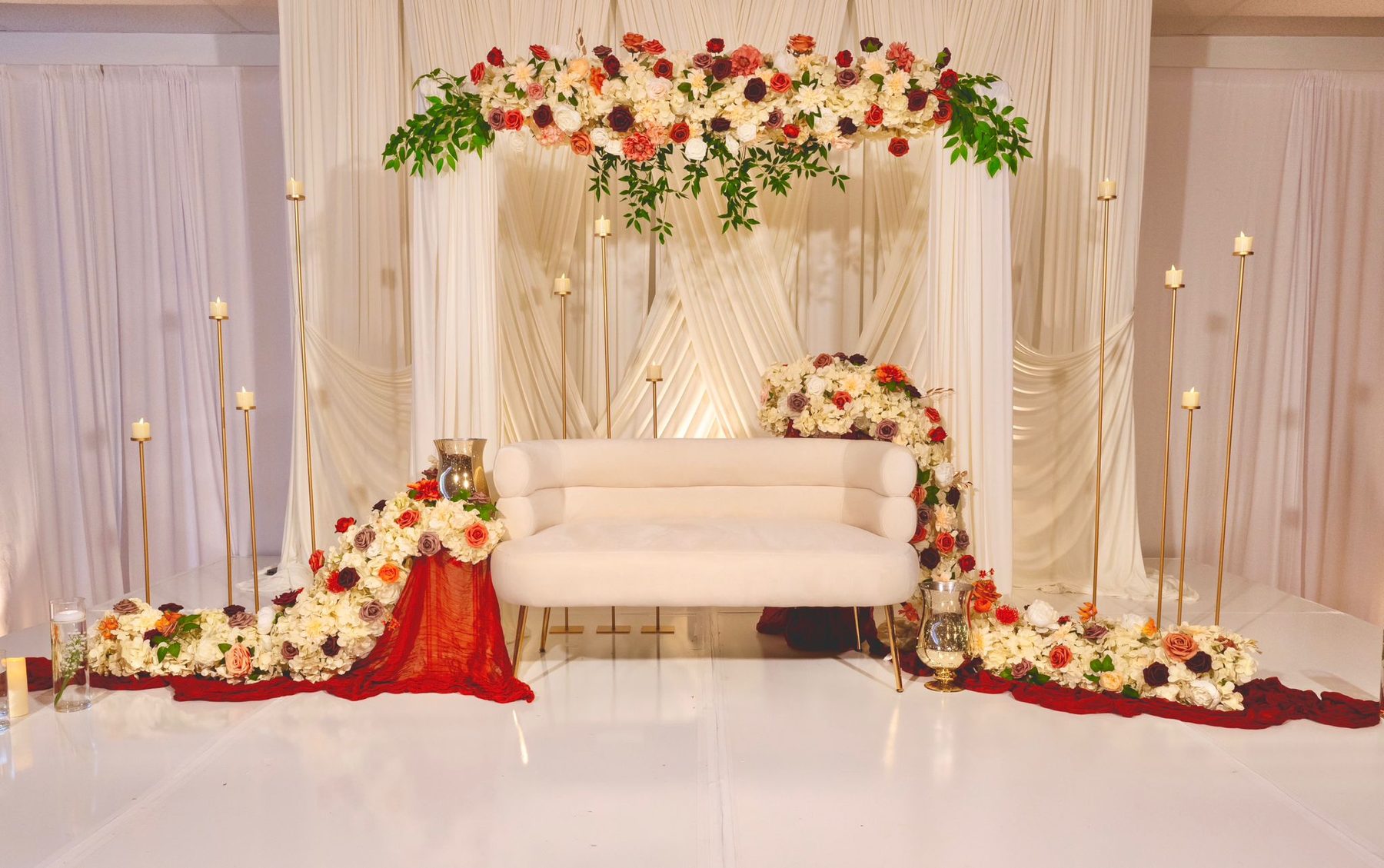 A floral sweetheart stage with an ivory loveseat and gold candle stands by Elysian Events