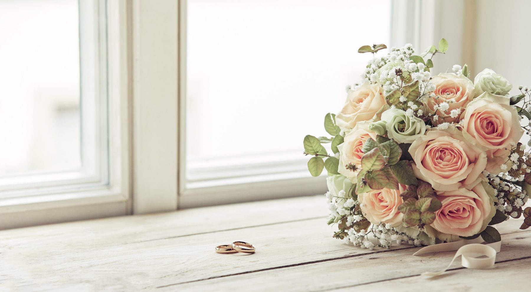 A bouquet of peach roses and baby&rsquo;s breath beside two gold wedding bands on a sunlit windowsill