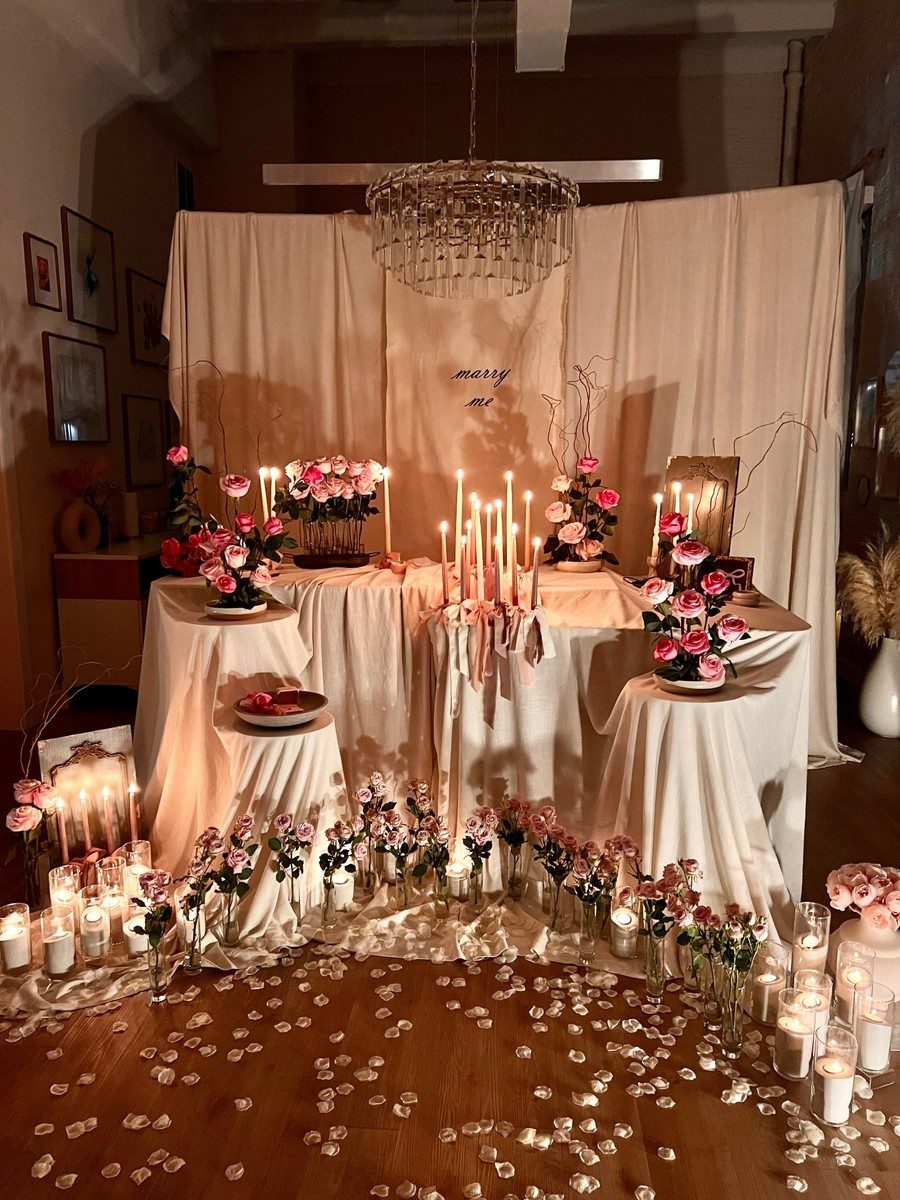 A candlelit proposal setup with draped ivory linens, pink roses and a &ldquo;marry me&rdquo; backdrop by bymayaht